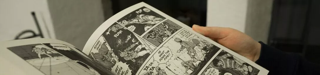 Close-up of a hand holding an open comic book with black and white artwork, showcasing detailed linework and shading.