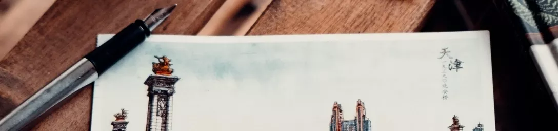 Close-up of a postcard lying on a wooden table, with a fountain pen placed beside it.