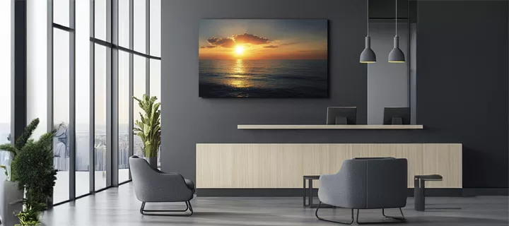 Modern lounge area with two gray chairs, a wooden reception desk, and a large wall-mounted sunset ocean photograph. Glass windows on the left offer natural light.