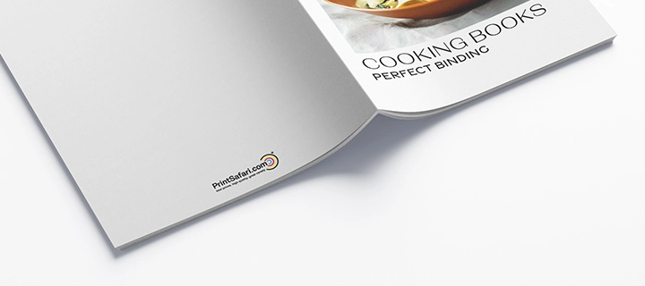 Cookbook lying open on a desk, with the cover facing up and pages spread out. The cover features a table setting with a meal and a glass of wine.