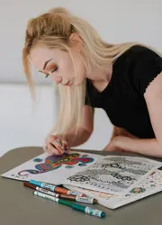 A woman focused on coloring detailed patterns in a coloring book, with a variety of colored pencils beside her.