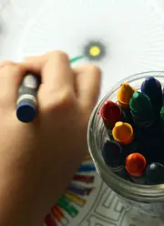 Top view of a hand holding a pen, coloring intricate designs in a coloring book, with a jar full of wax pens positioned beside the hand.